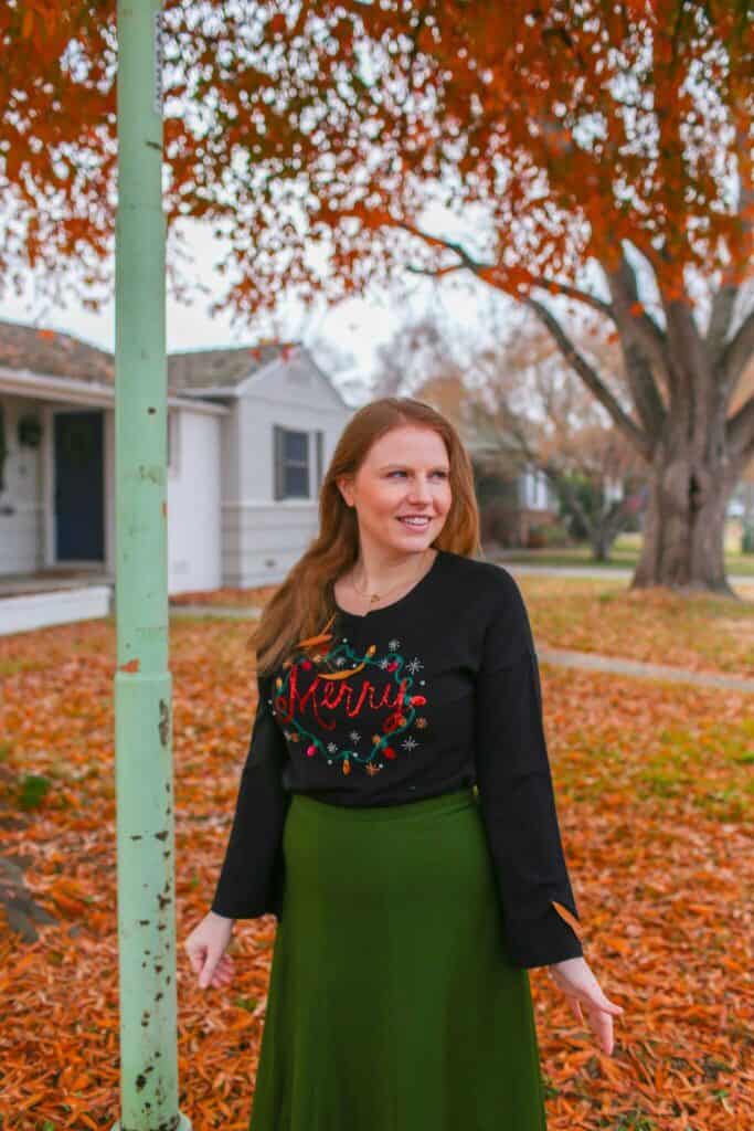 Festive woman wearing Christmas sweater outdoors during autumn foliage, celebrating holiday season.