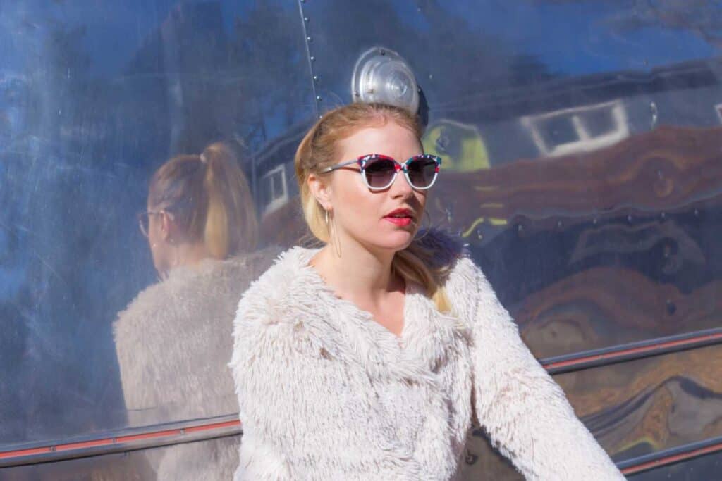 Vibrant woman wearing colorful sunglasses and cozy sweater, sitting outdoors against a reflective metallic background.