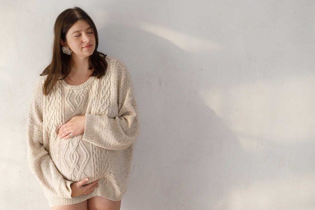 Woman wearing a cozy cream sweater posing indoors for soft and minimalist winter maternity photos.