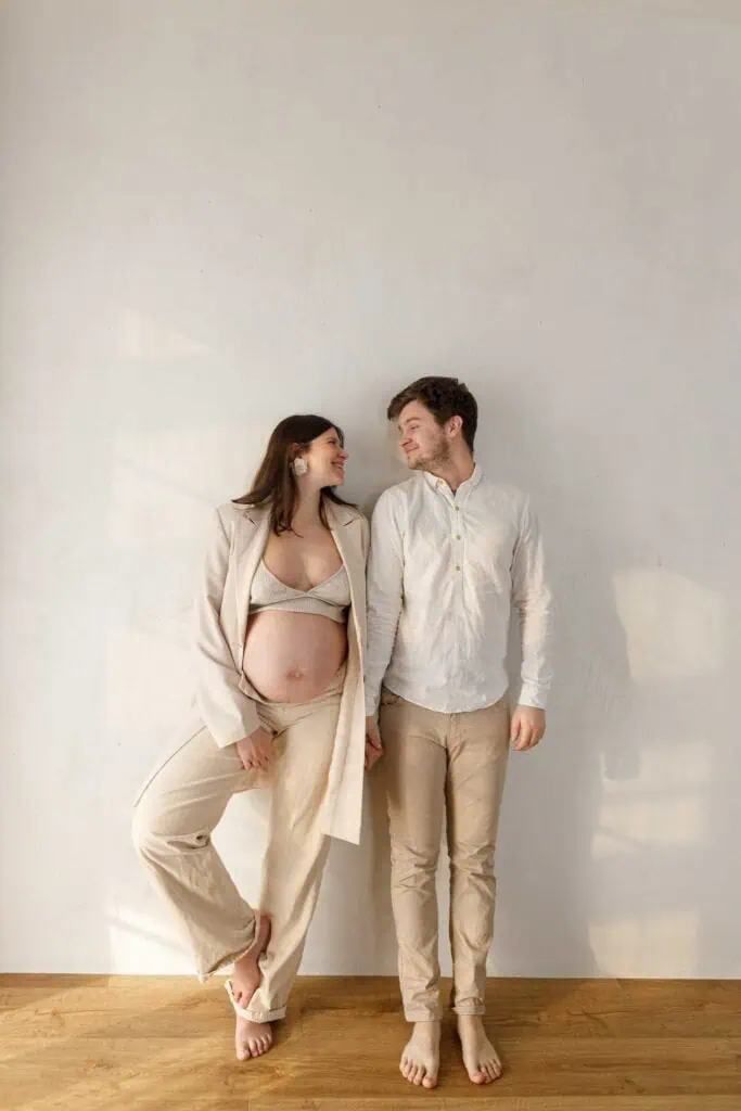 Expecting couple smiling at each other during couples maternity photos by a minimalist wall.